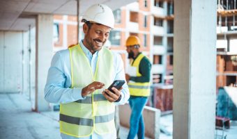 Shot of a young businessman using his smartphone while on a construction site. Portrait of young engineer in vest with white helmet standing on construction site, smiling and holding smartphone for worker, internet, social media.