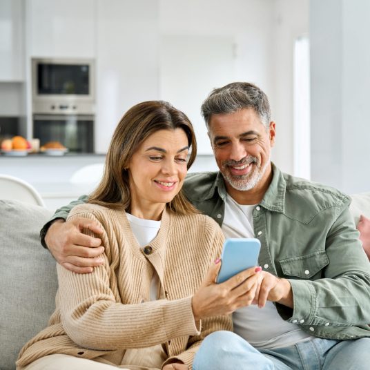 Happy older mature senior family couple, middle aged man and woman looking at cell phone using smartphone mobile technology device together sitting at home relaxing on couch doing online shopping.